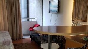 A compact hotel room with a bed, a small dining table with two stools, a TV mounted on the wall, and a window with curtains.