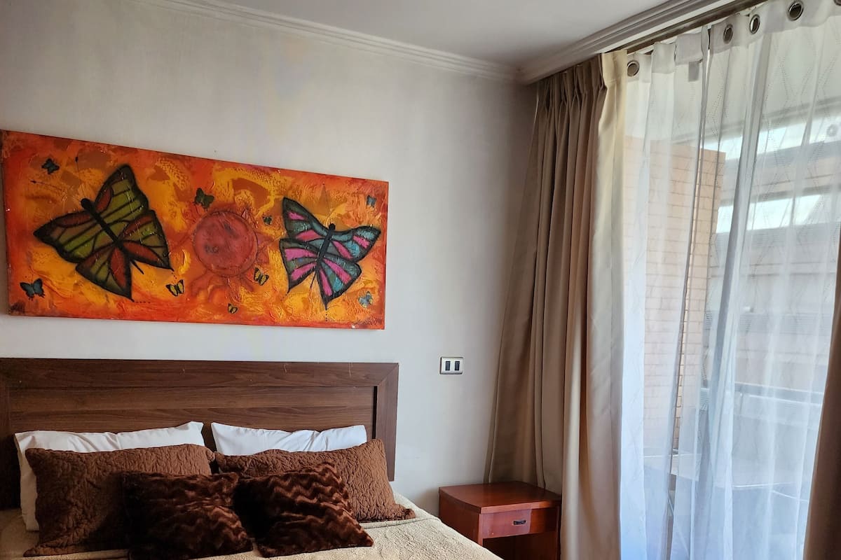 Un dormitorio con una cama grande, una mesita de noche, un cuadro y una ventana con cortinas.
