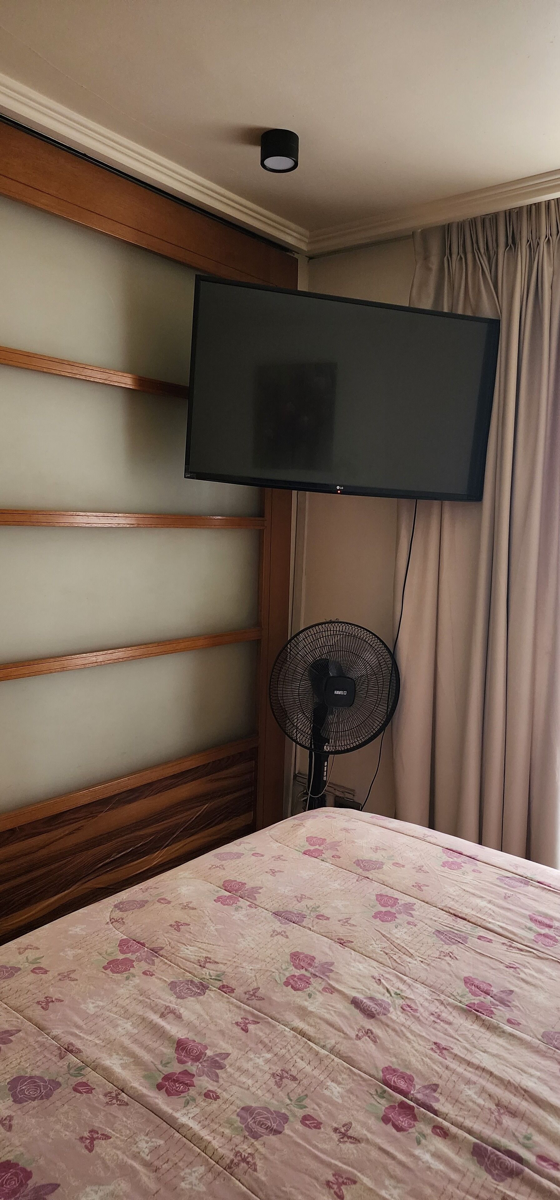 A hotel room with a bed, a flat-screen TV mounted on the wall, a standing fan, and a window with curtains.