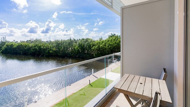 A balcony with a glass railing overlooking a river, a wooden deck, and a view of a lush green landscape under a clear sky. - Luxury condo within the Seven Mile Beach Corridor (Grand Cayman)