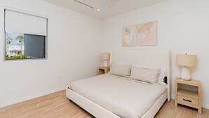 A modern bedroom with a bed, two bedside tables, a ceiling fan, a window with a view, and two wall art pieces. - Luxury condo within the Seven Mile Beach Corridor (Grand Cayman)