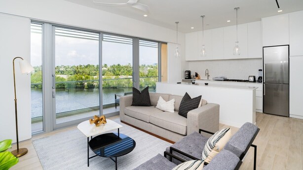 Un appartement moderne comprenant un espace de vie, une cuisine et de grandes portes-fenêtres coulissantes donnant sur un balcon avec vue sur la verdure et l’eau.