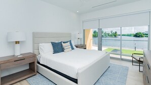 A modern bedroom with a large bed, a nightstand, a lamp, and a view of a body of water through large windows. - Luxury 2 Bed 2 Bath Premium Condo - (Grand Cayman)