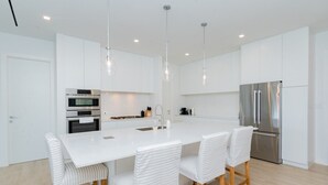 A modern kitchen with white cabinetry, stainless steel appliances, and a central island with seating. - Luxury 2 Bed 2 Bath Premium Condo - (Grand Cayman)