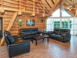 A log cabin interior with a wooden ceiling, leather sofas, a wooden coffee table, and a large window with a view of greenery.