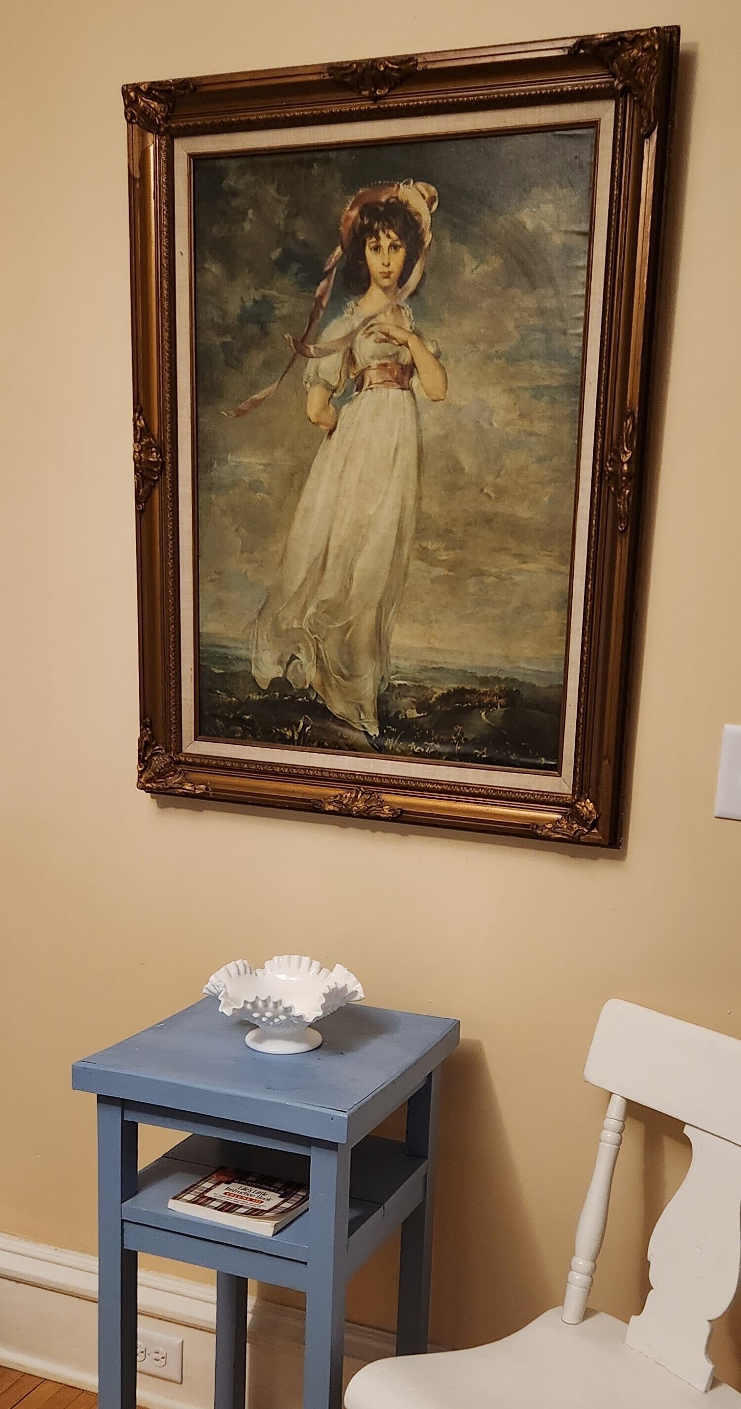 A framed painting on a wall, a small blue table with a book, and a white chair.