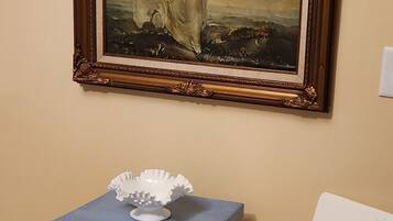 A framed painting on a wall, a small blue table with a book, and a white chair.