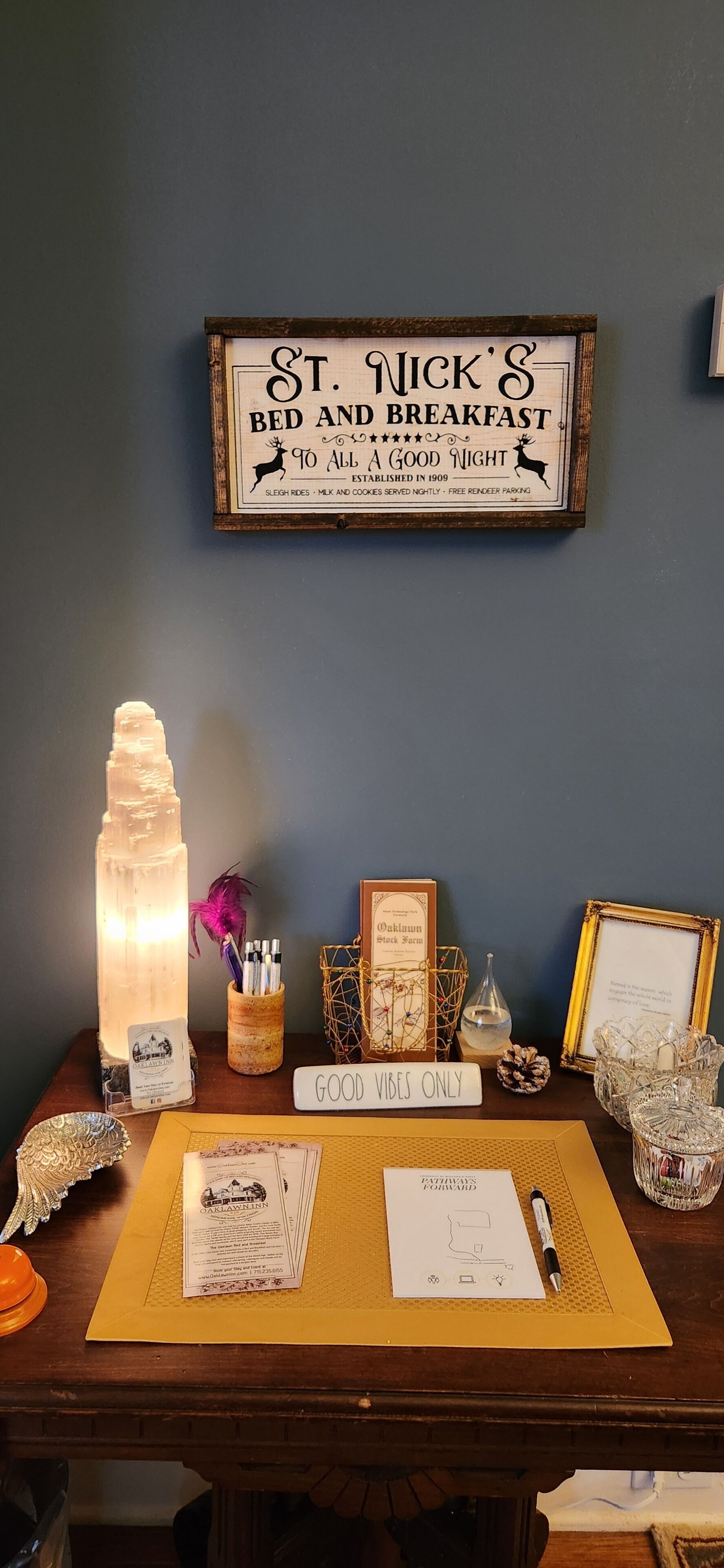 Un escritorio con un cartel enmarcado que dice "St. Nick's Bed and Breakfast", una lámpara, un cartel que dice "Solo buenas vibras" y varios objetos decorativos.