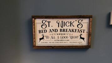 Un escritorio con un cartel enmarcado que dice "St. Nick's Bed and Breakfast", una lámpara, un cartel que dice "Solo buenas vibras" y varios objetos decorativos.
