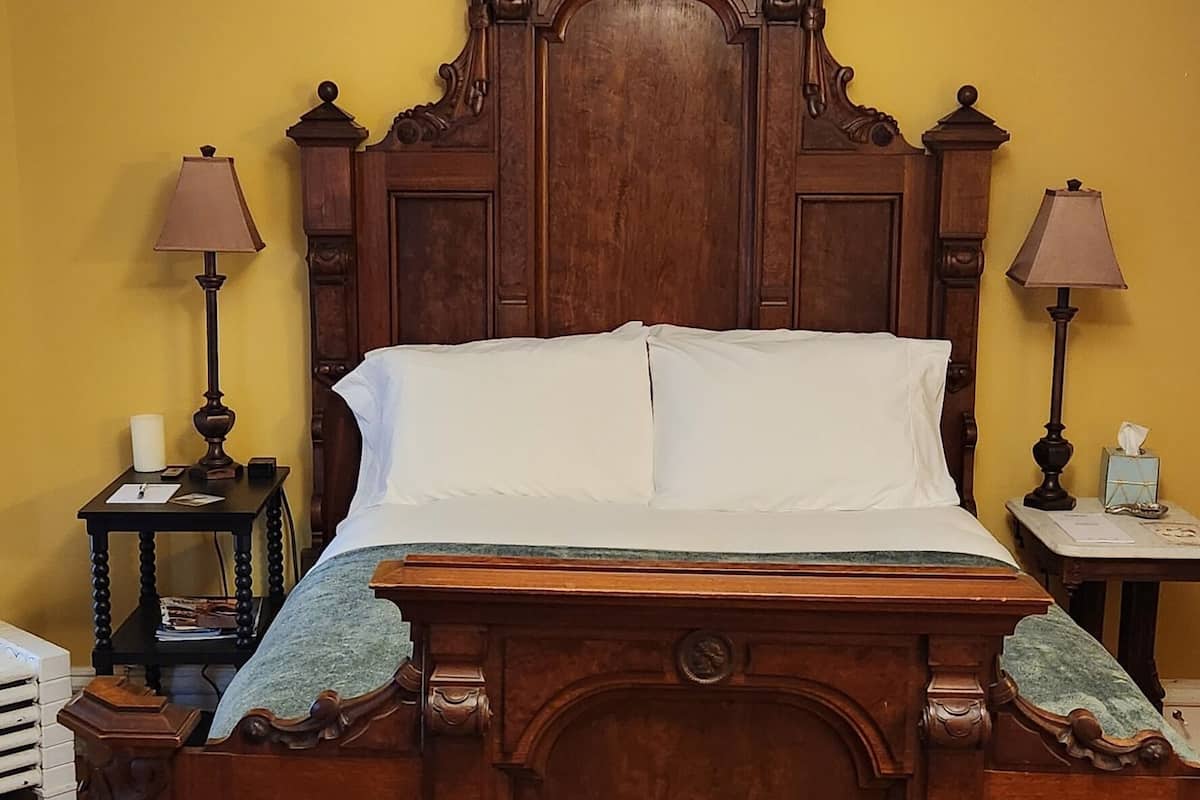 A traditional wooden four-poster bed with a matching headboard, flanked by two bedside tables with lamps, in a room with yellow walls and wooden flooring.
