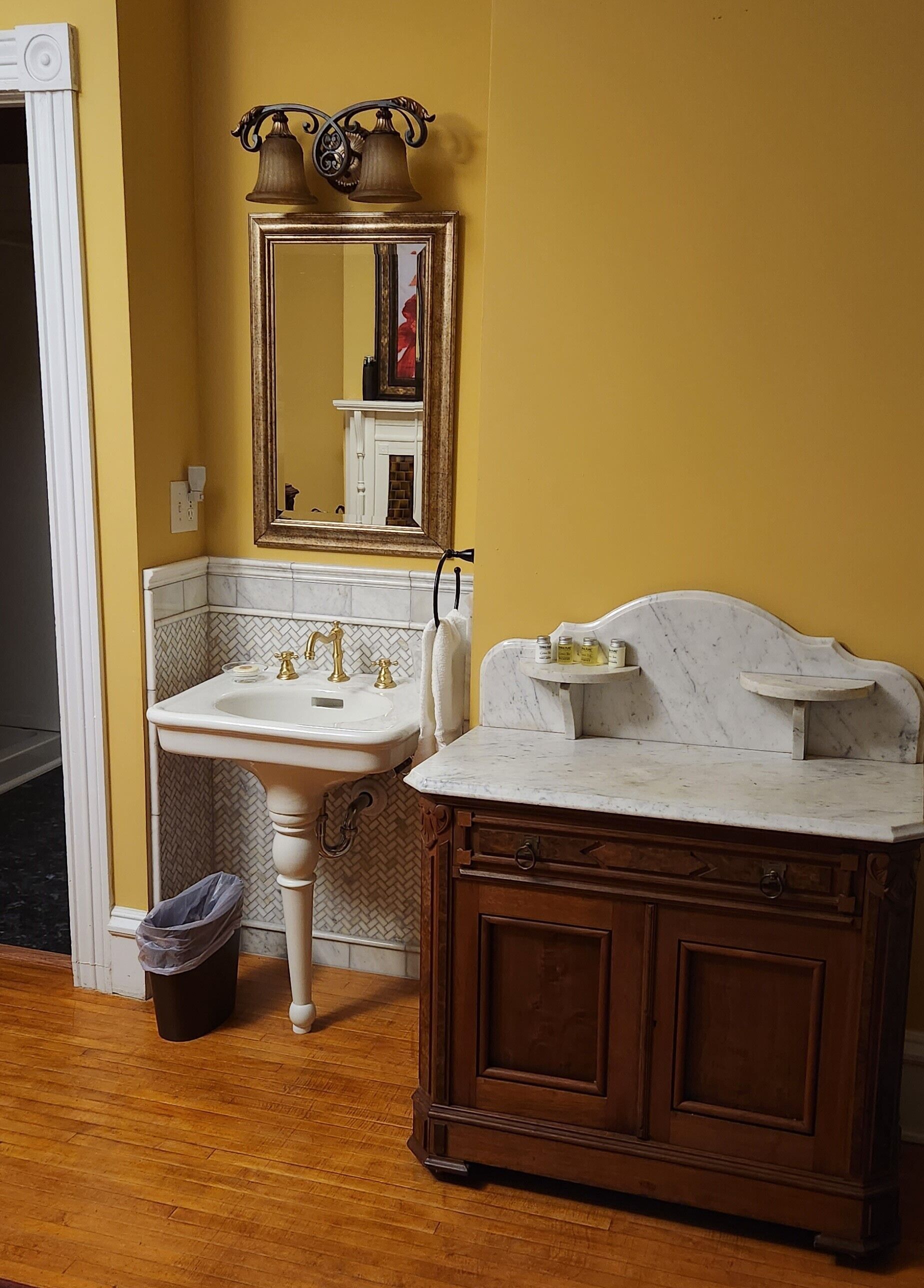 Une salle de bain avec un lavabo sur colonne blanc, un meuble-lavabo en bois, un miroir avec un cadre décoratif et une poubelle.