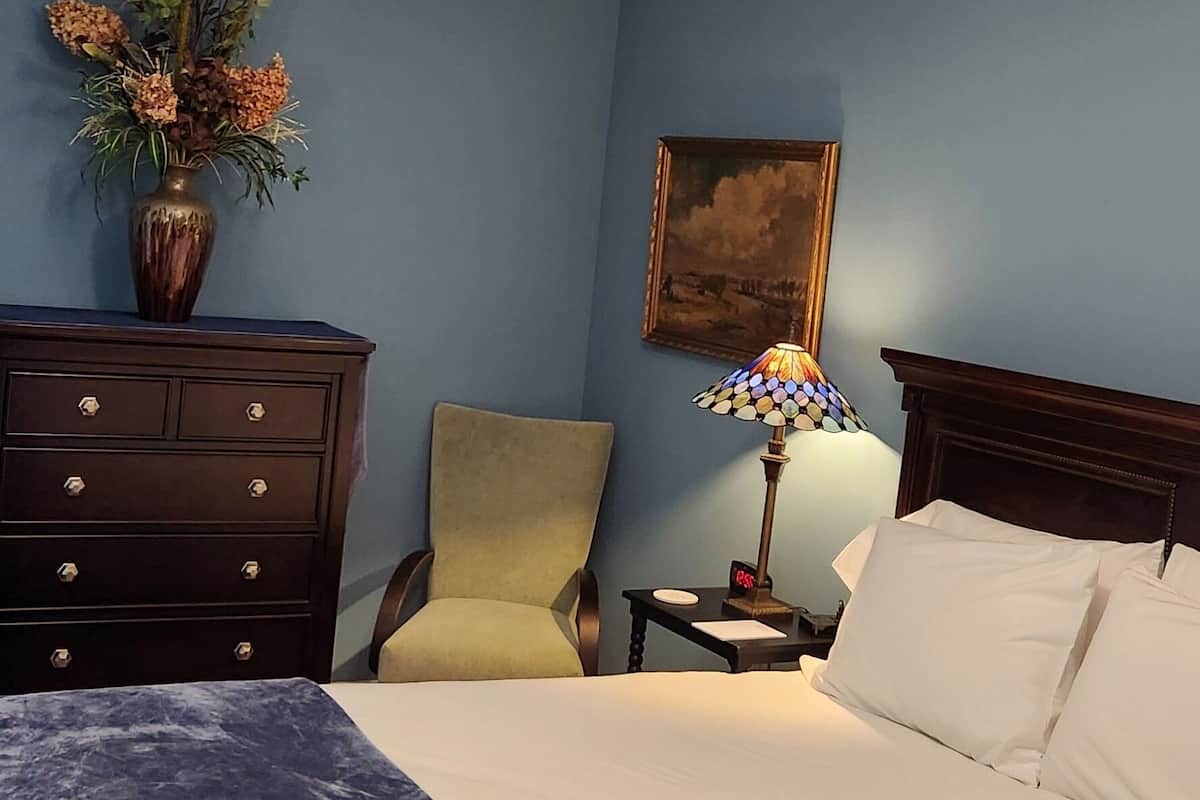 A bedroom with a bed, a nightstand, a lamp, a chair, a dresser, and a vase with flowers.