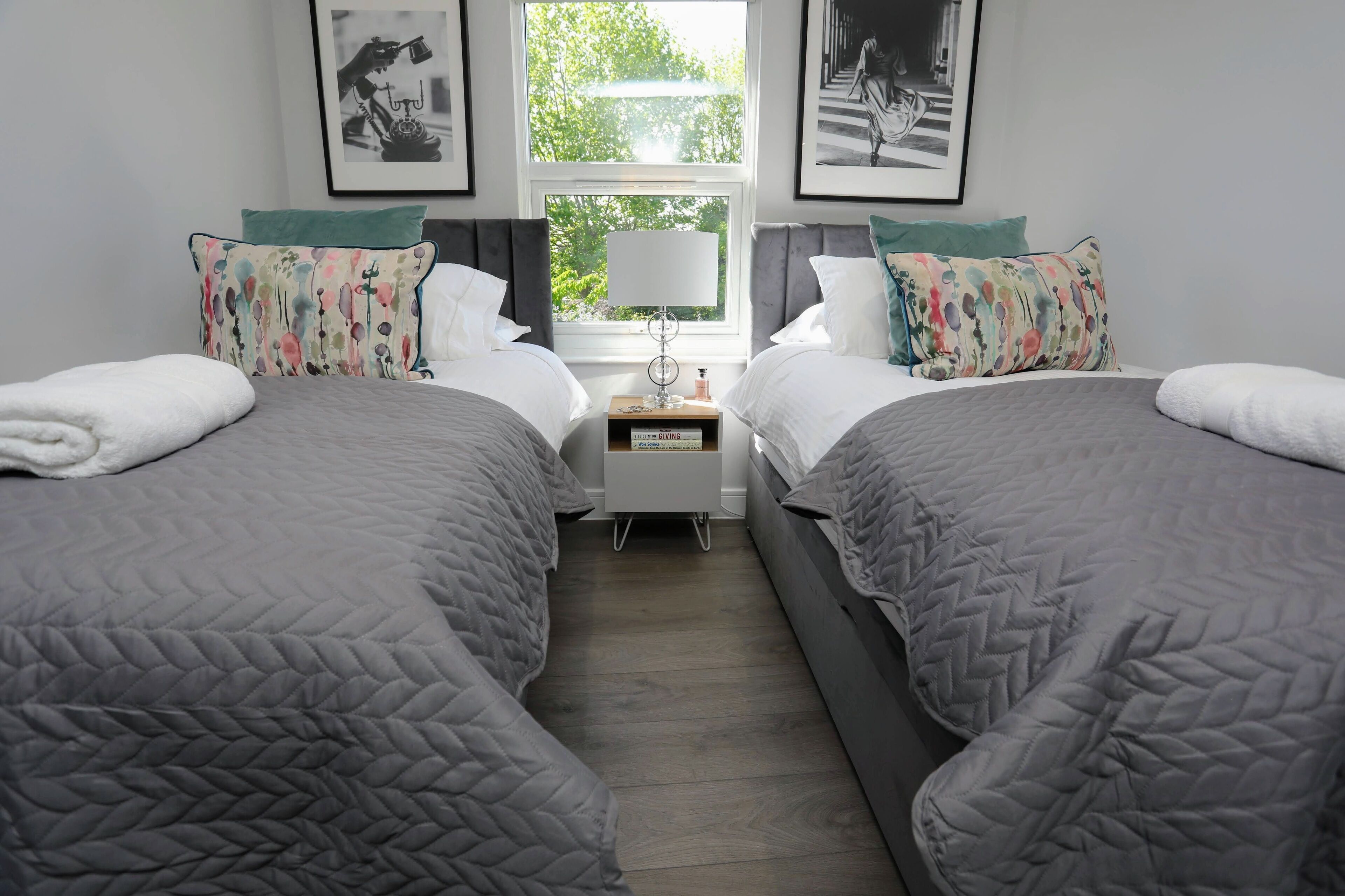 A twin bedroom with two beds, each with a grey quilted duvet, a bedside table with a lamp, and a small wooden nightstand.