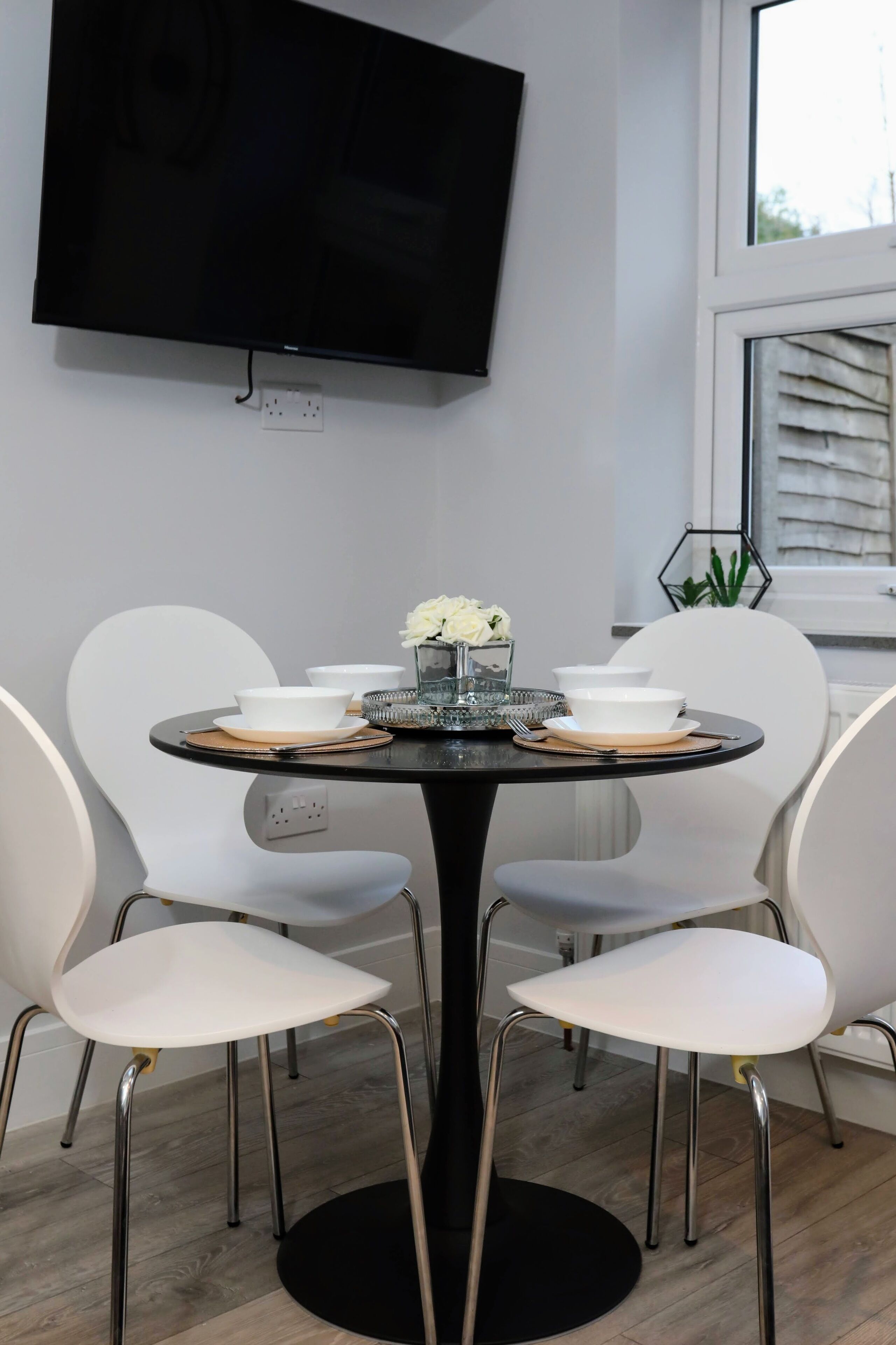 Un comedor con una mesa redonda, sillas blancas y un televisor negro montado en la pared.