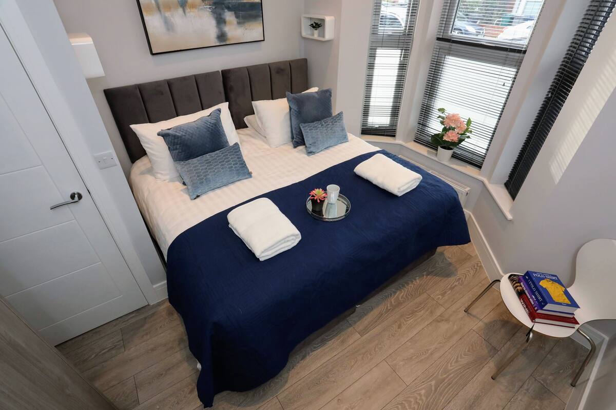 A neatly made bed with a blue bedspread and white pillows, a bedside table with a book and a glass, and a window with blinds and a flower vase.