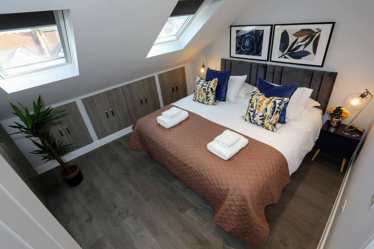 A bedroom with a sloped ceiling, a large bed with a brown quilt, multiple pillows, a nightstand with a lamp, and a potted plant.