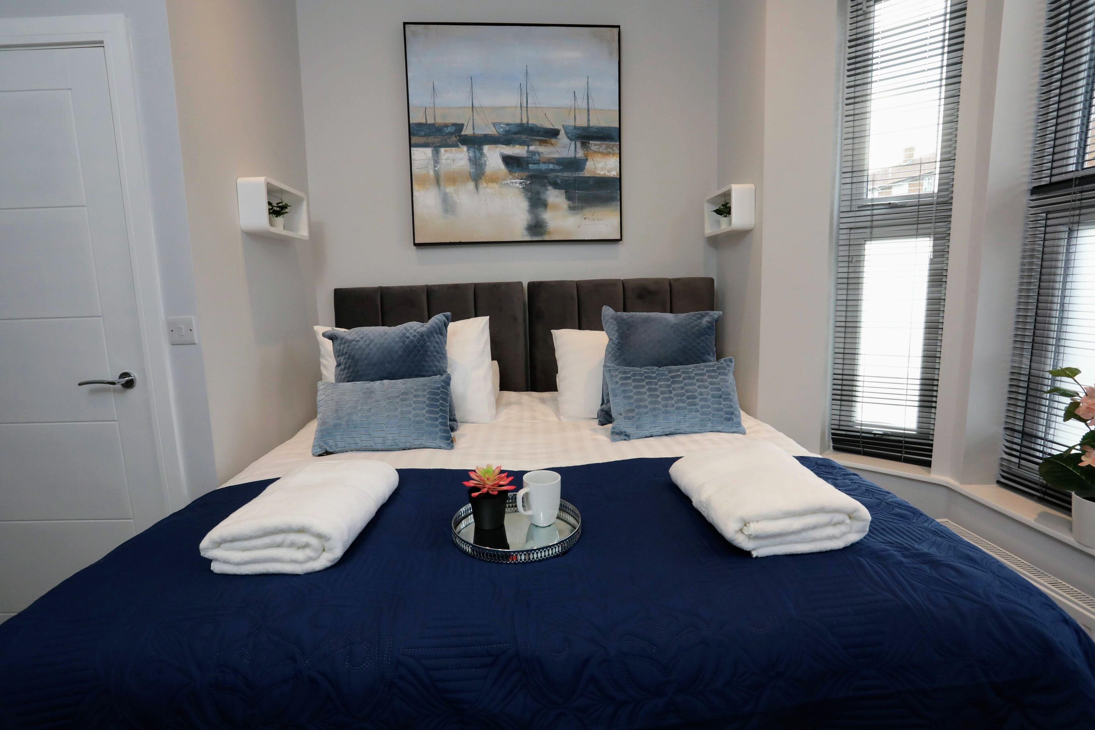 A neatly made bed with a blue bedspread, two white towels, and a tray with a mug and a flower.