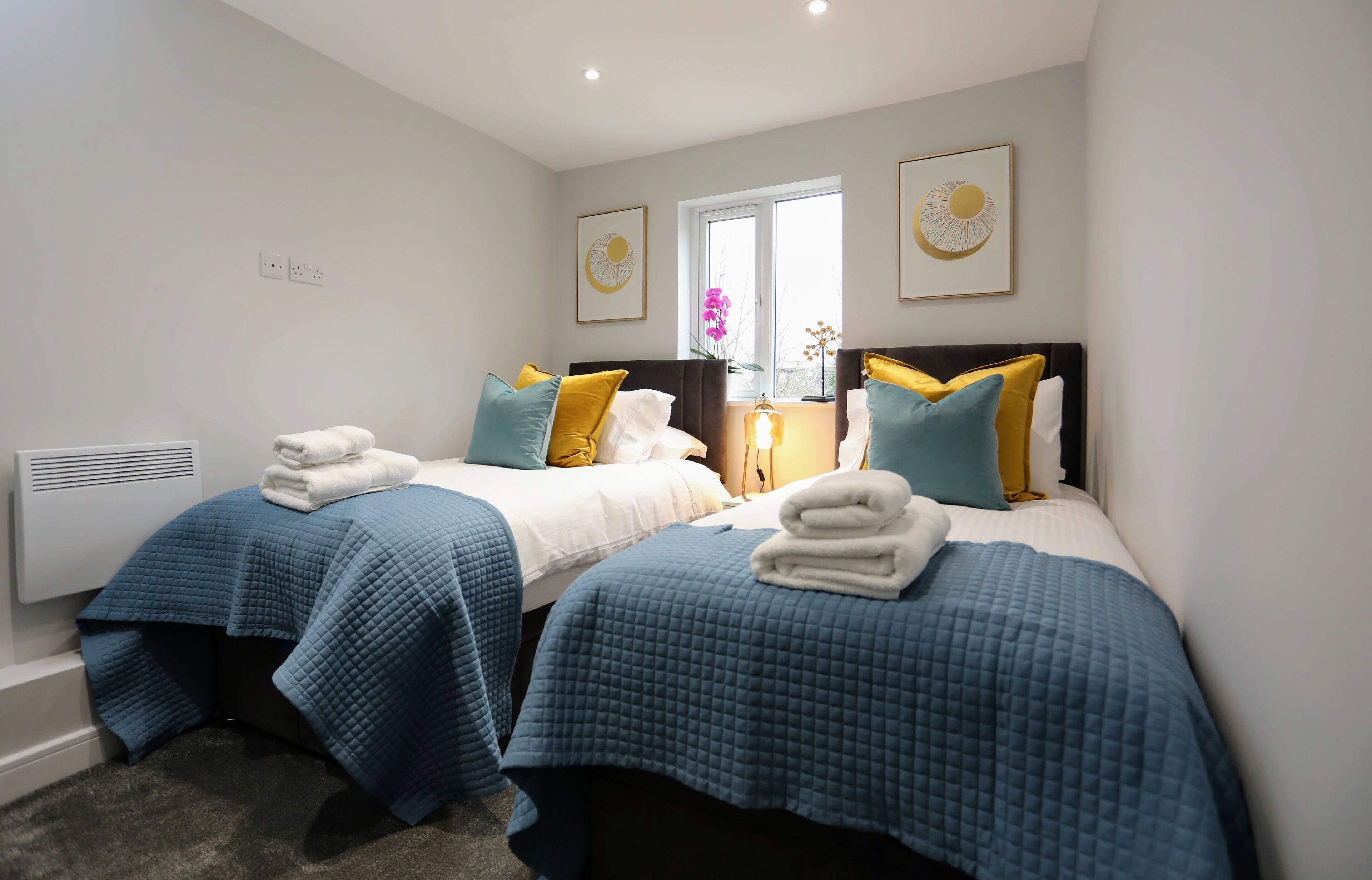 A hotel room with two beds, each with blue bedding and white towels. There are two wall-mounted light fixtures and a window with a view of the outdoors.