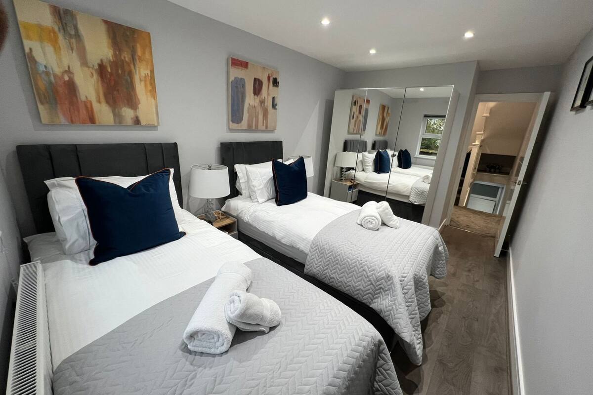 A hotel room with two beds, white bedding, and blue pillows. There are two paintings on the wall, a mirror, and a door leading to a bathroom.