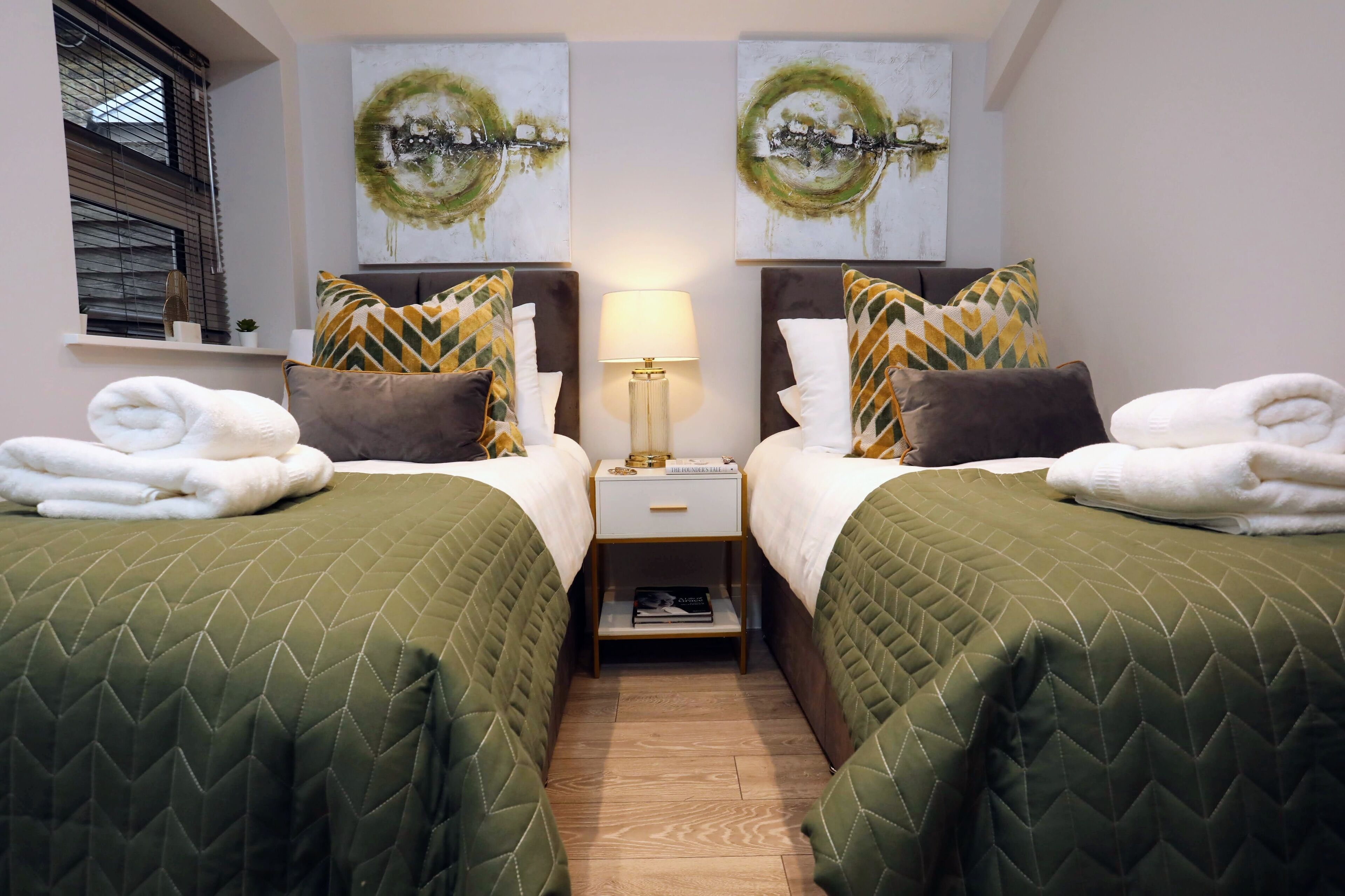 A hotel room with two beds, each with green quilts and patterned pillows, a bedside table with a lamp, and artwork above the beds.