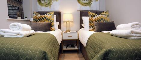 A hotel room with two beds, each with green quilts and patterned pillows, a bedside table with a lamp, and artwork above the beds.