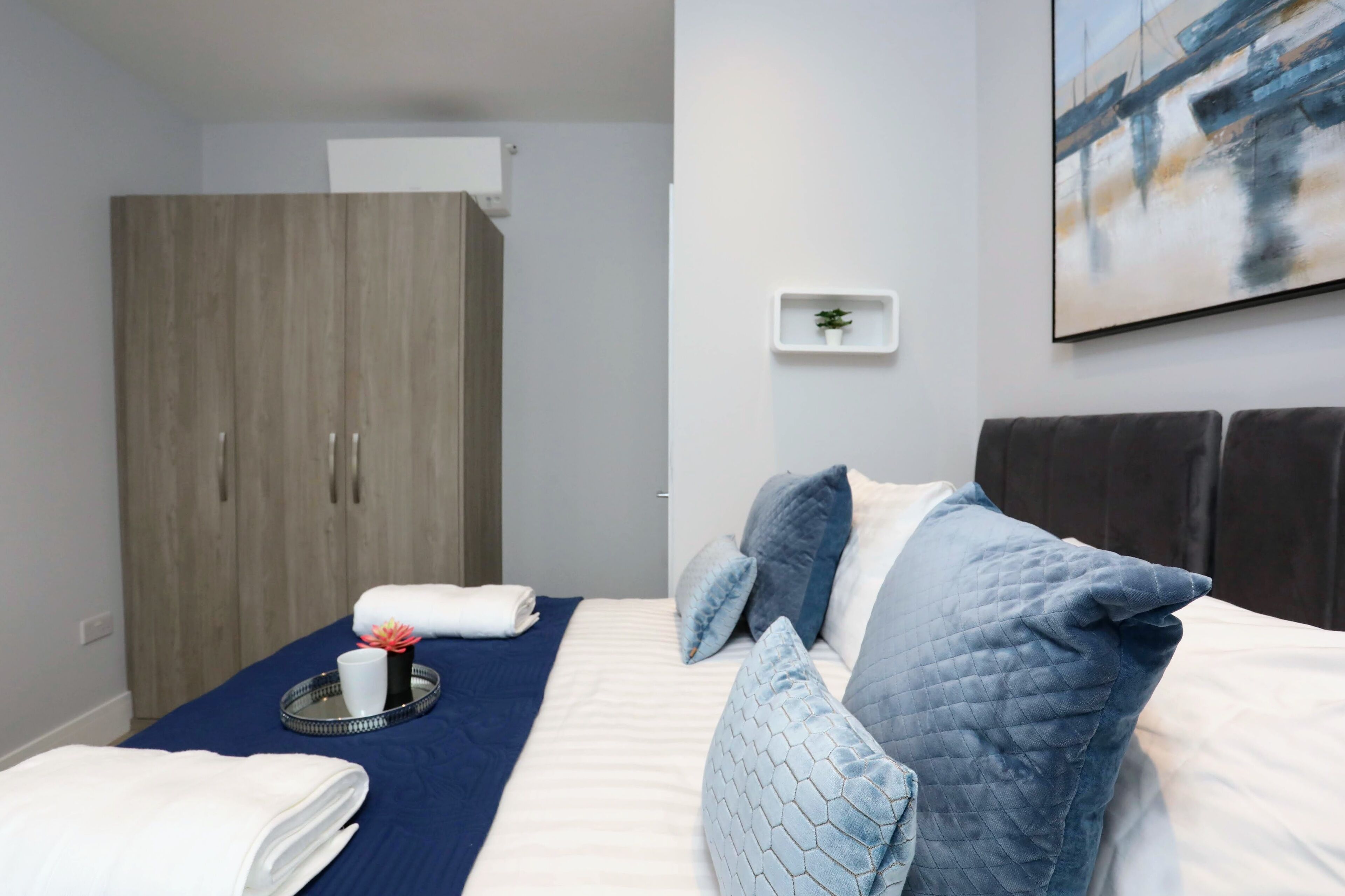 A hotel room with a bed, a wardrobe, a small shelf with a plant, and a painting on the wall.
