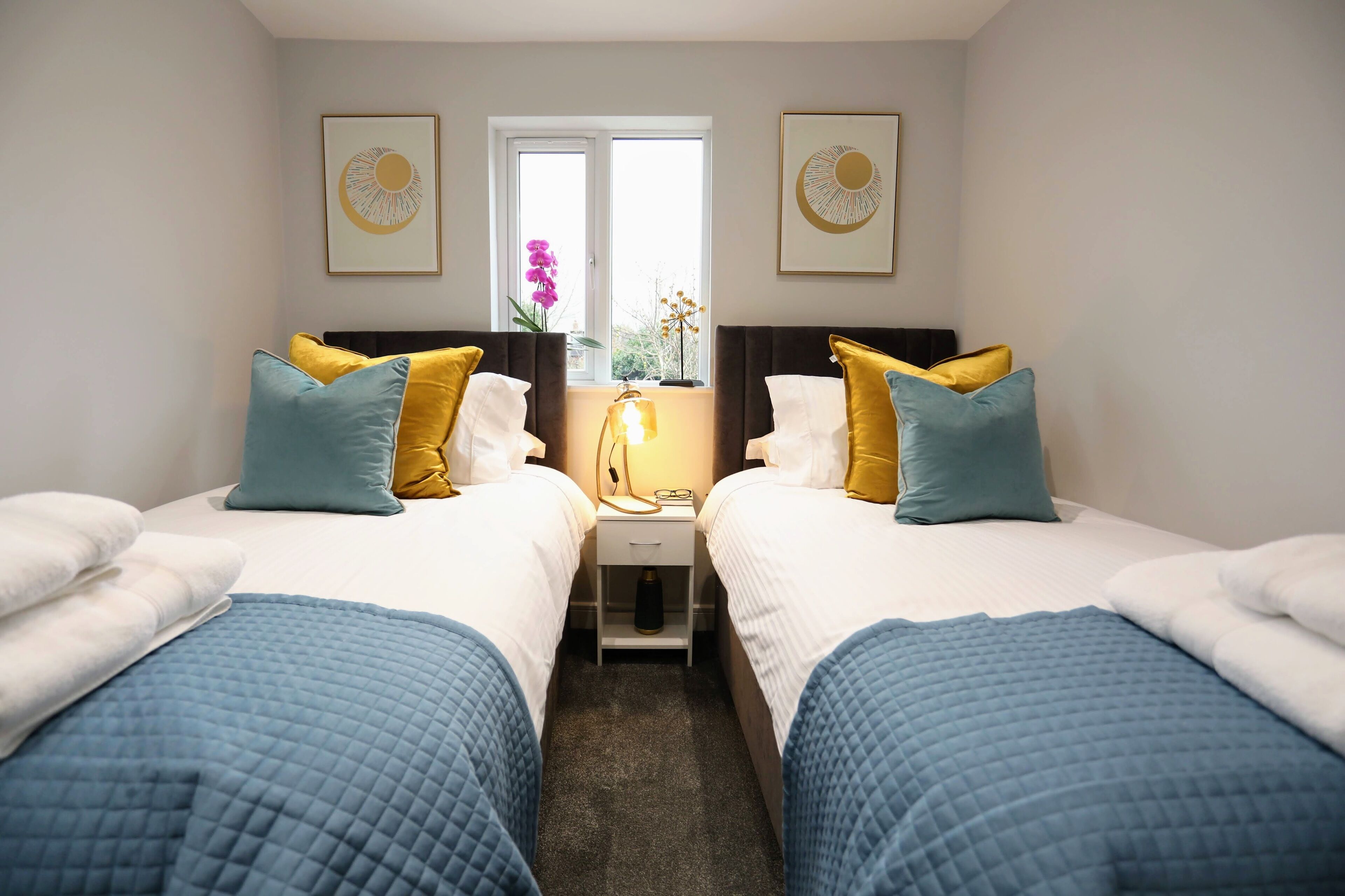 A hotel room with two beds, each with a blue quilted bedspread and pillows in yellow and blue. There is a nightstand with a lamp between the beds, and two framed pictures on the walls.
