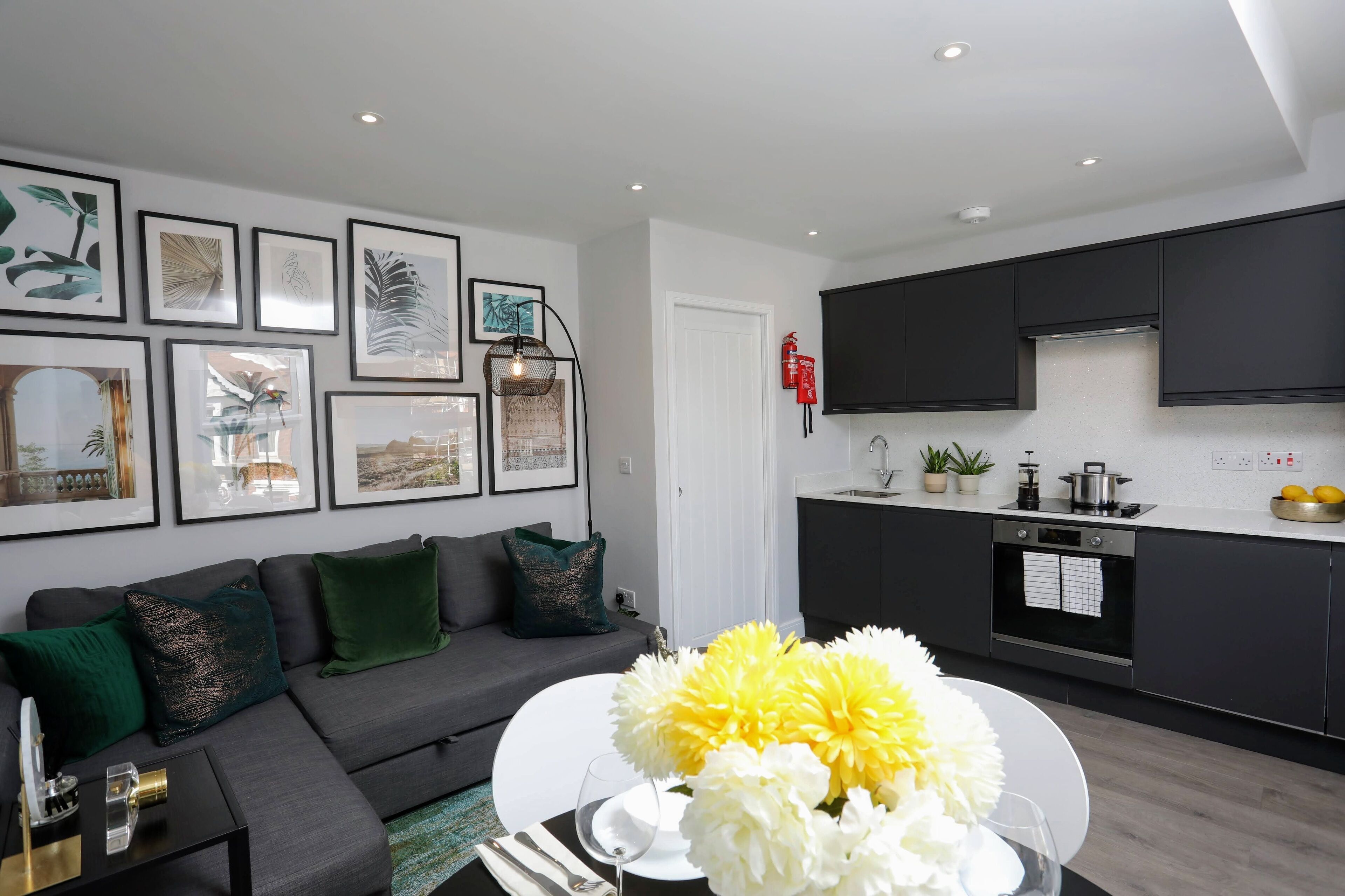 En moderne stue med en grå sofa, grønne puder og et spisebord med hvide blomster. Et køkken med mørke skabe og en hvid bordplade ses i baggrunden.
