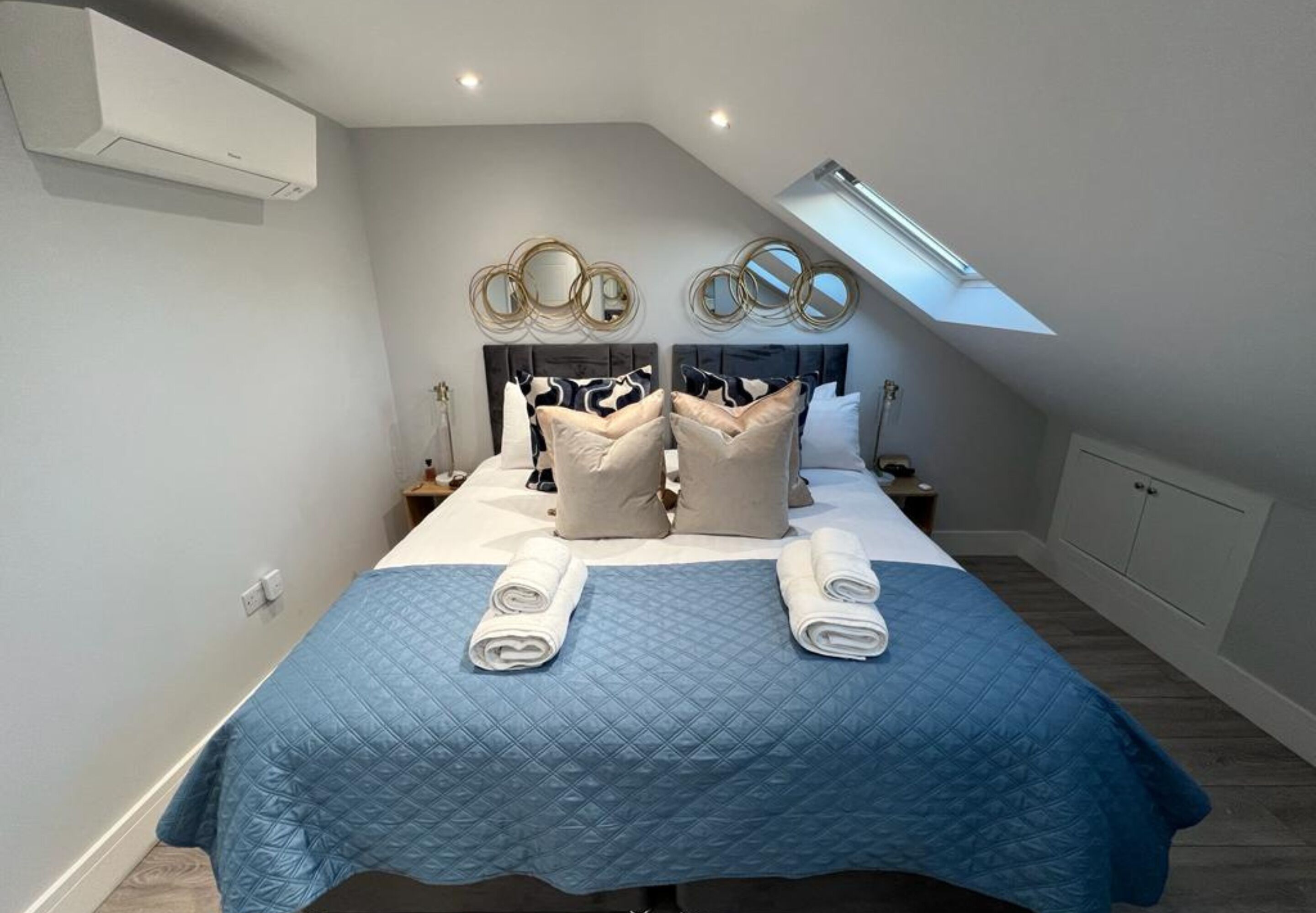 A bedroom with a bed, two bedside tables, a skylight, and a wall-mounted air conditioner.
