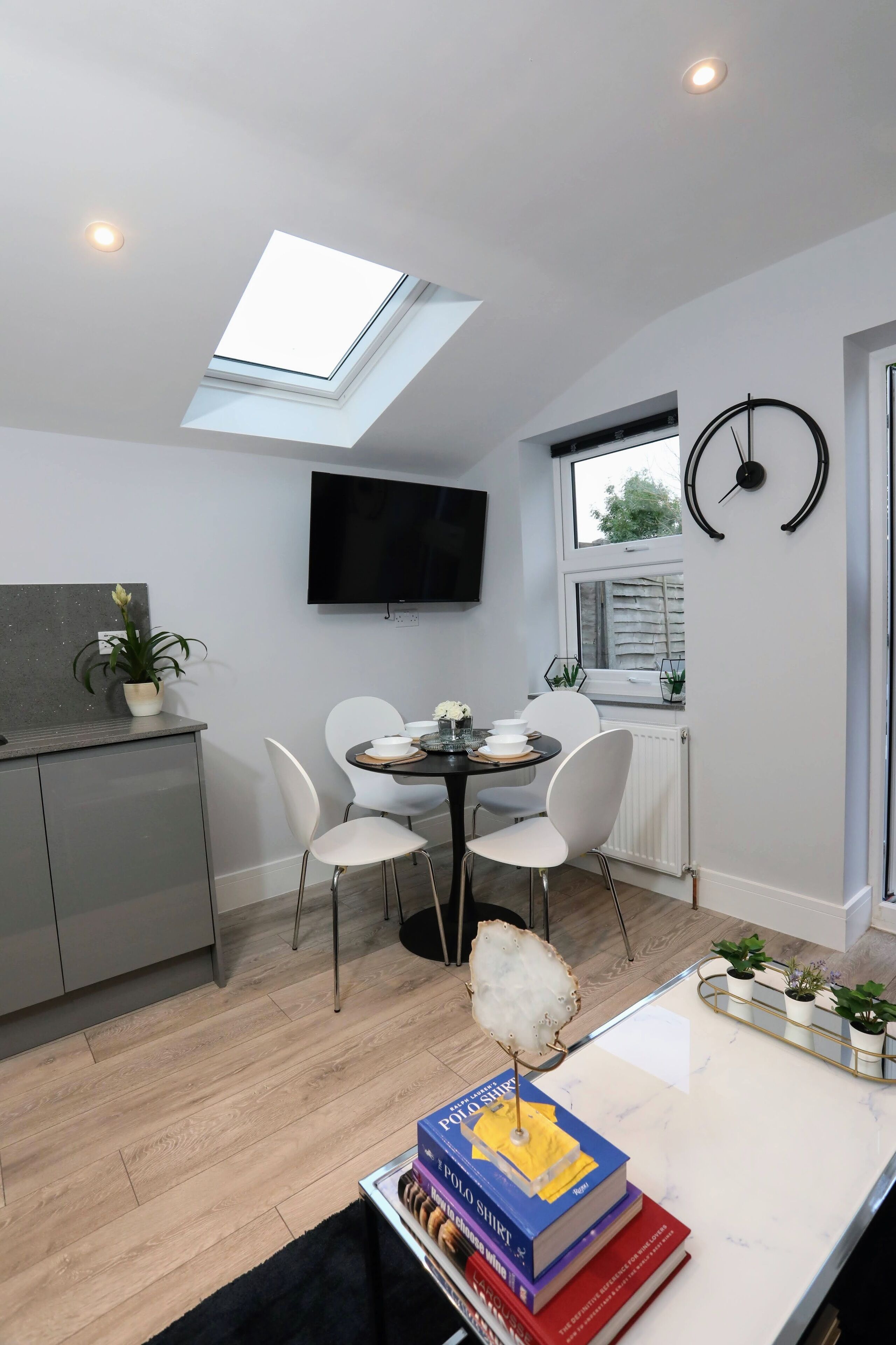 A compact kitchen with a skylight, a dining area with white chairs, a TV mounted on the wall, and a window with blinds.
