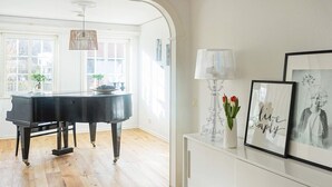 Une pièce spacieuse avec un grand piano, un parquet en bois, une console blanche ornée d’œuvres d’art et une grande fenêtre laissant entrer la lumière naturelle.