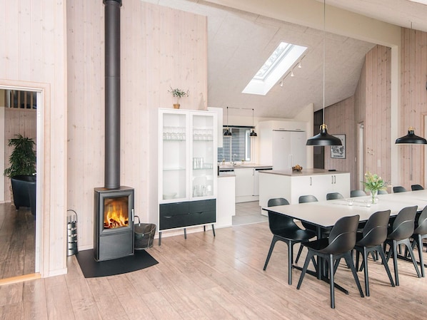A modern kitchen with a central island, a dining area with a long table and chairs, and a visible wood-burning stove.