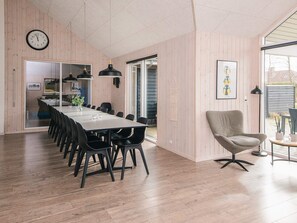 Un espace repas moderne avec une longue table, des chaises noires, des luminaires suspendus et une horloge au mur.