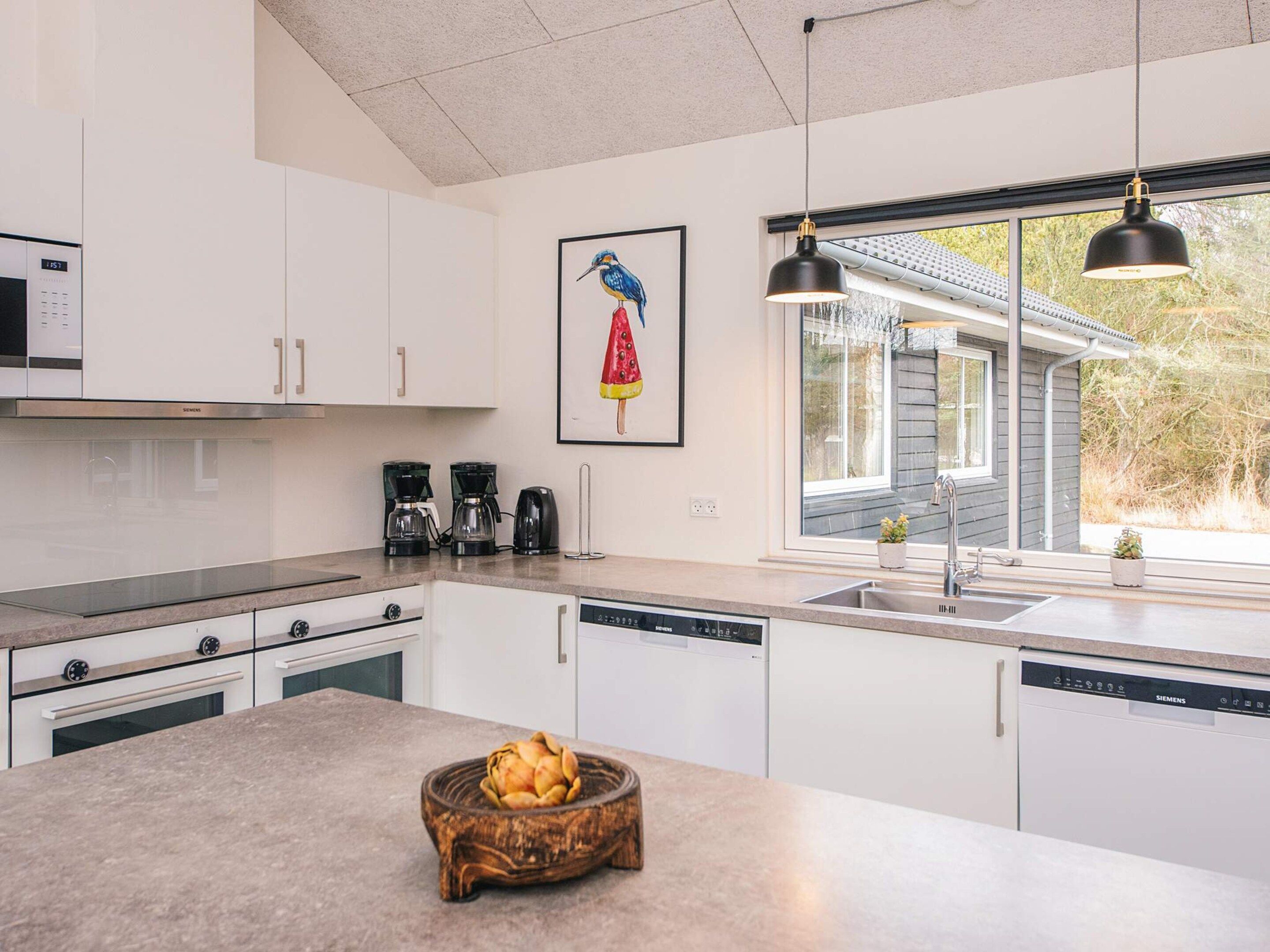 Una cocina moderna con armarios blancos, una isla grande, una estación de café y una ventana con vista a una casa y árboles.
