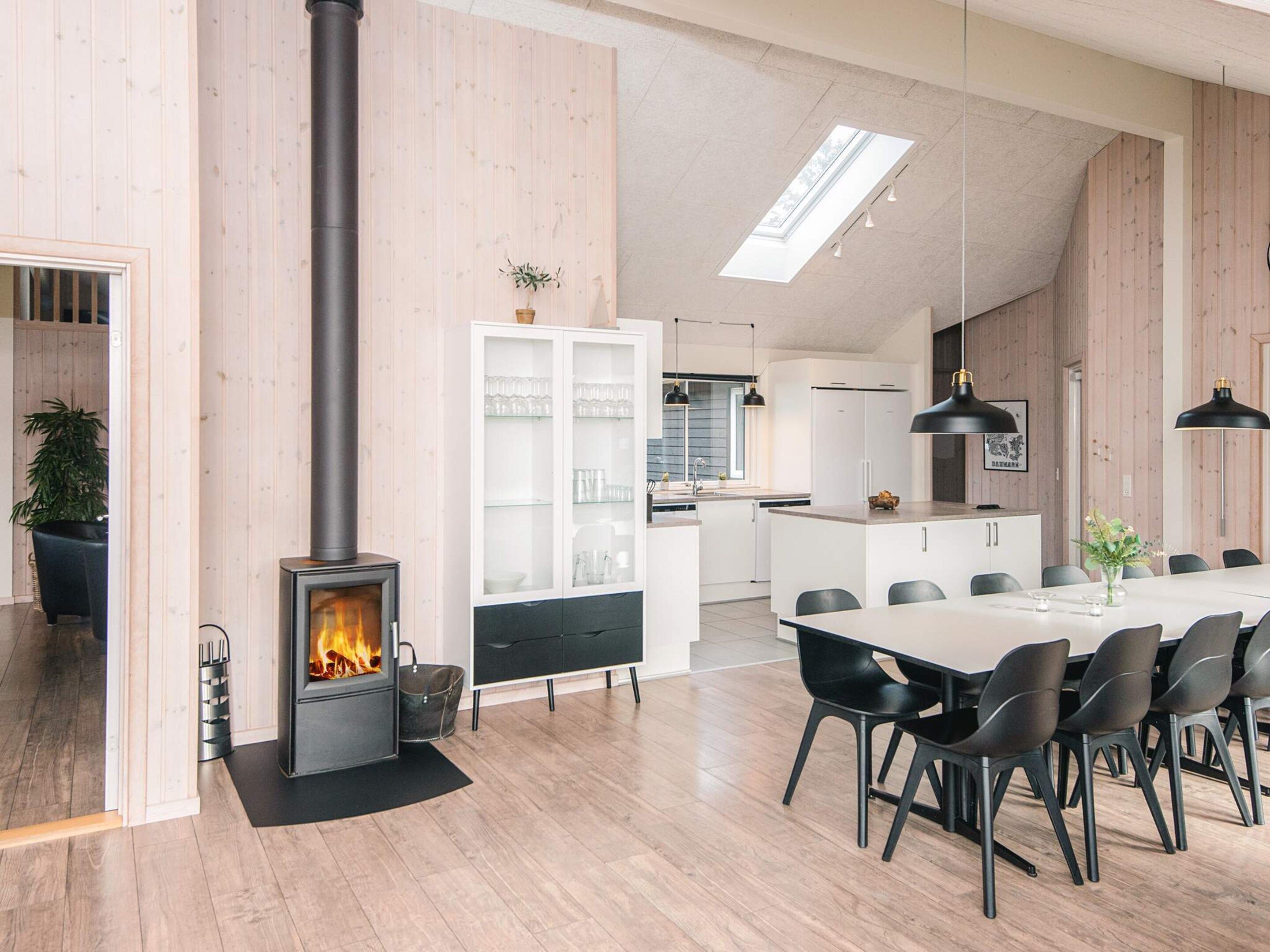 Una cocina moderna con una isla central, una zona de comedor con una mesa larga y sillas, y una estufa de leña visible.