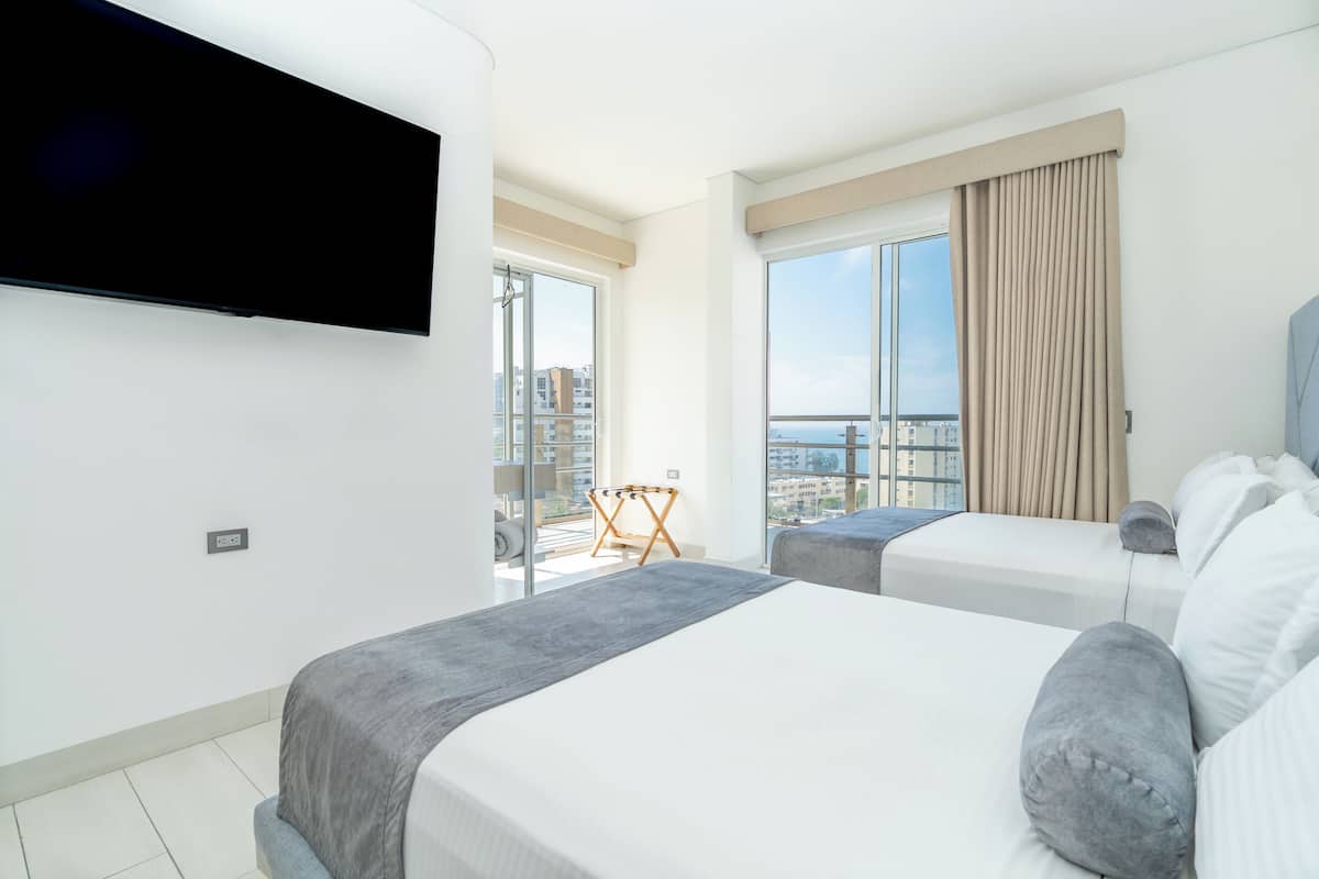 A hotel room with two beds, a flat-screen TV mounted on the wall, and a balcony with a view of the city.