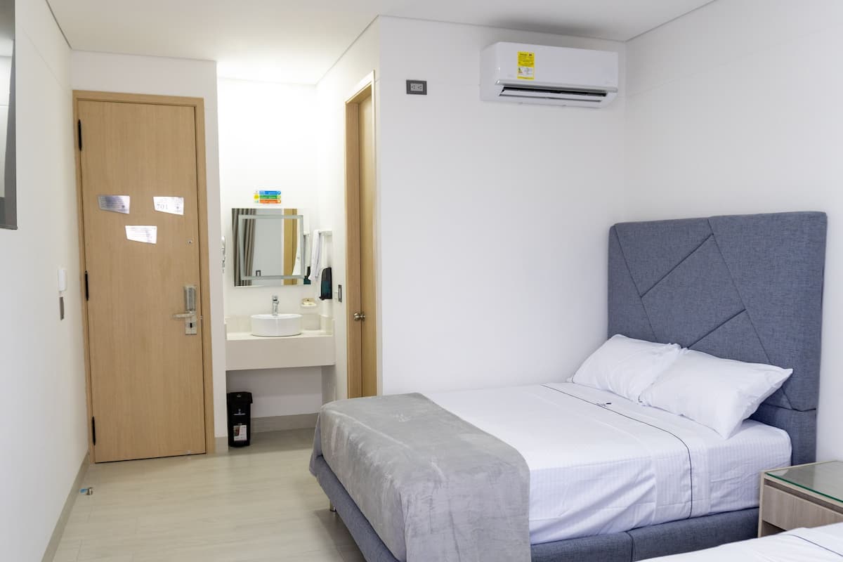 A bedroom with a bed, a bedside table, a mirror, a bathroom with a sink and mirror, and an air conditioning unit.