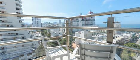 A balcony with two white chairs and a view of a cityscape and the sea.