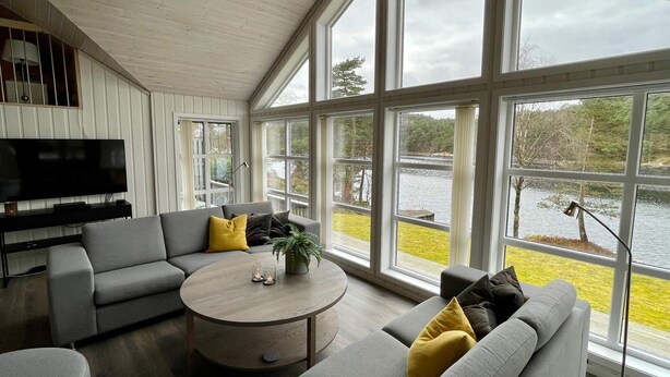 Un salon moderne avec un canapé gris, une table basse ronde et un téléviseur à écran plat fixé au mur. De grandes fenêtres offrent une vue sur un lac et la végétation environnante.