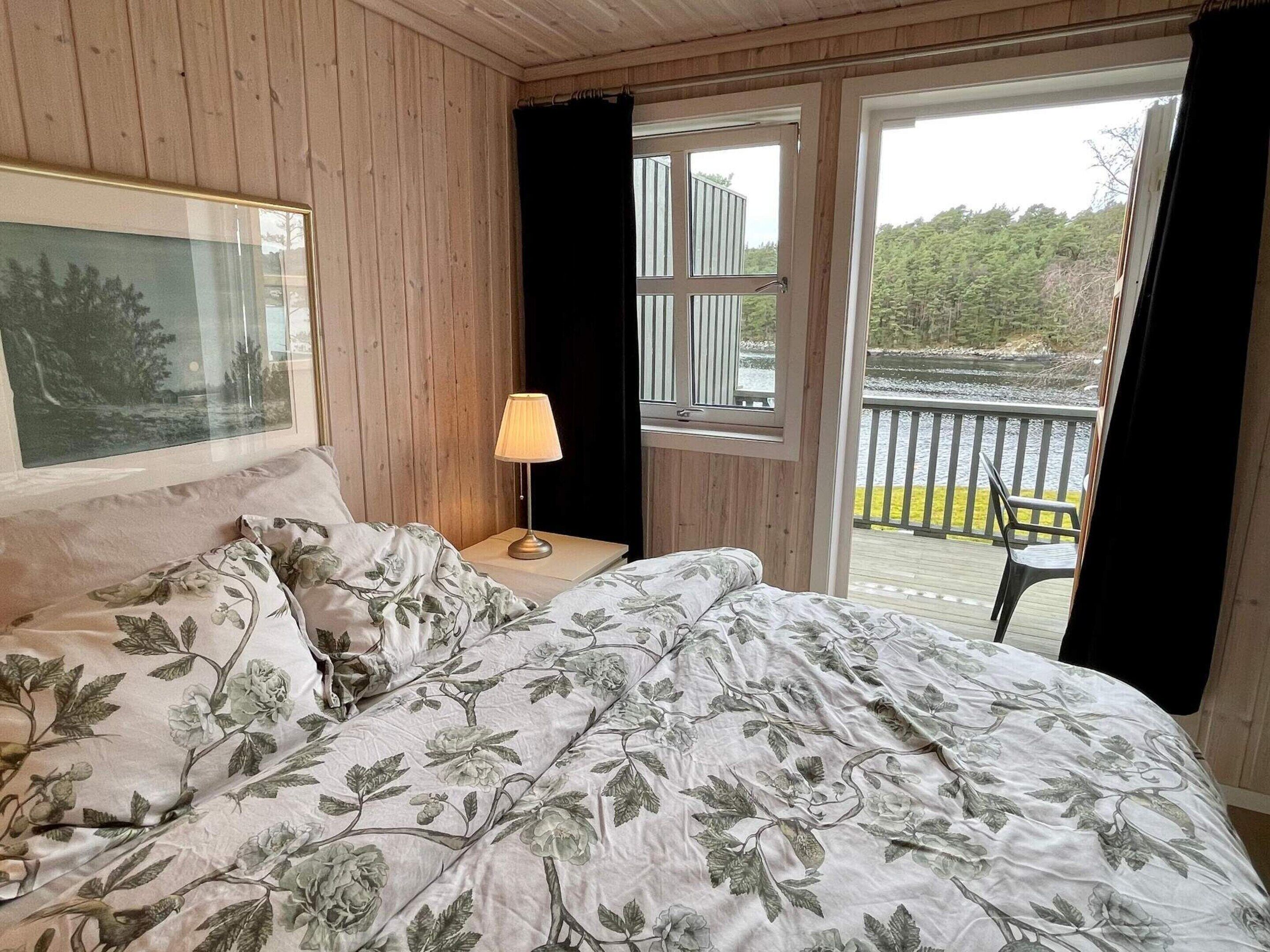 Un dormitorio con cama, mesita de noche, lámpara, ventana y vistas a una terraza y árboles.