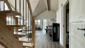 Un intérieur spacieux avec un escalier en bois menant à une mezzanine, un coin repas et une cuisine dotée d’un poêle noir.