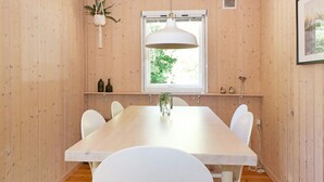 Een eetruimte met een houten tafel en witte stoelen, een hanglamp, een raam met uitzicht op groen, en een plank met decoratieve objecten.