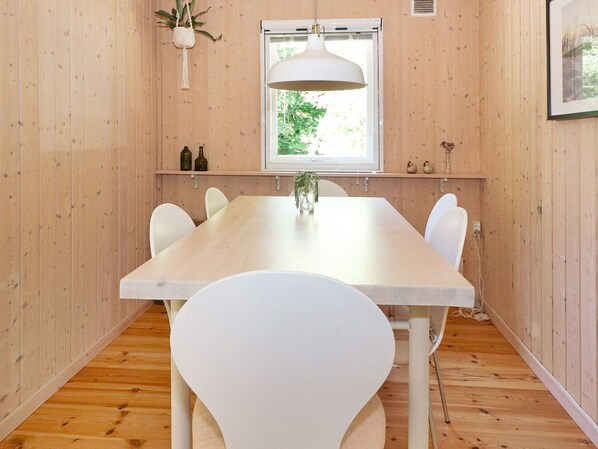 A dining area with a wooden table and white chairs, a hanging light fixture, a window with a view of greenery, and a shelf with decorative items.
