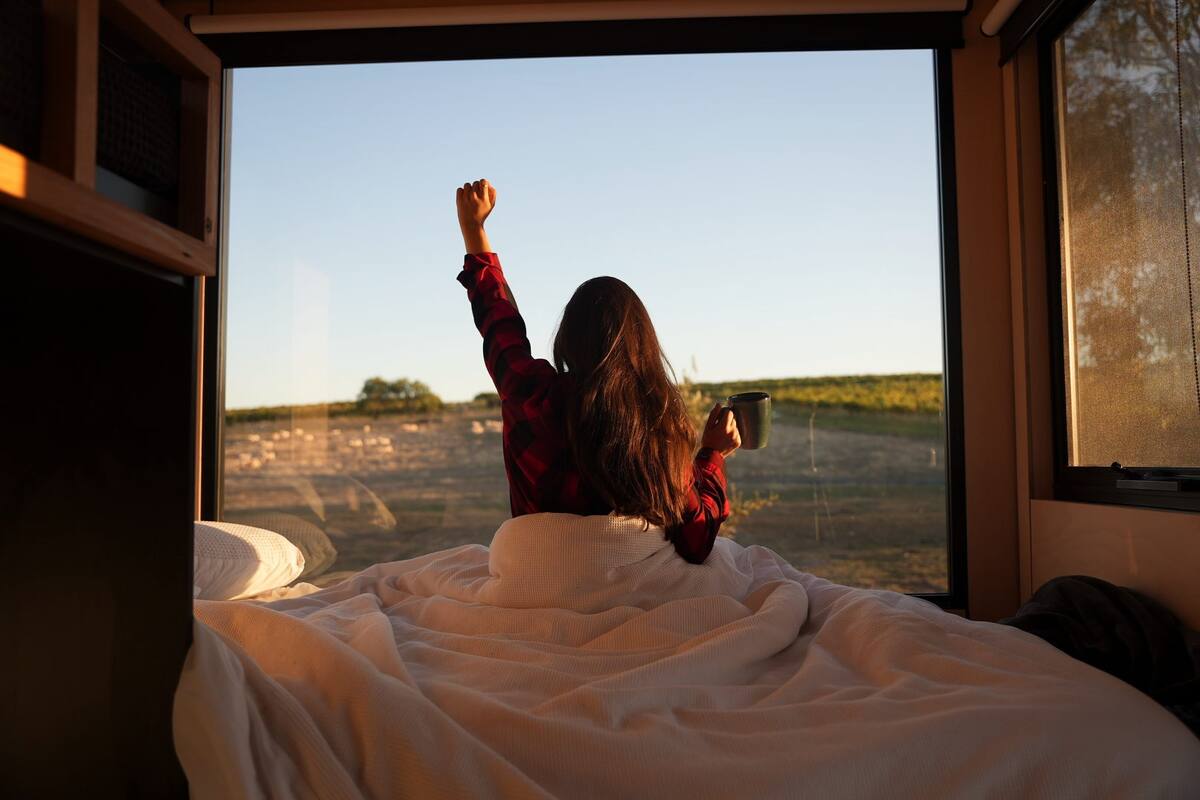 A bed with a white blanket, a person sitting on it, and a window with a view of a field.