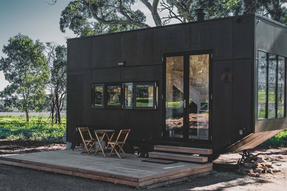 A modern, compact cabin with a deck, outdoor seating, and large windows offering views of a natural landscape.