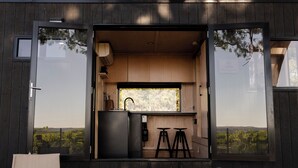 A compact, modern kitchen with a large window, a refrigerator, and two stools. - CABN McLaren Vale (McLaren Flat)