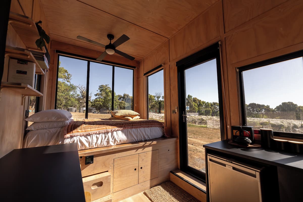A compact, wooden interior with a bed, a desk, and large windows offering a view of the outdoors.