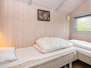 A single bed with a white mattress and pillows, a wooden wall, a framed picture, and a window with a view of trees.