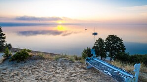 A bench on a sandy beach with a calm lake and trees in the background. - 5 Star Holiday Home in Borkop (Borkop)