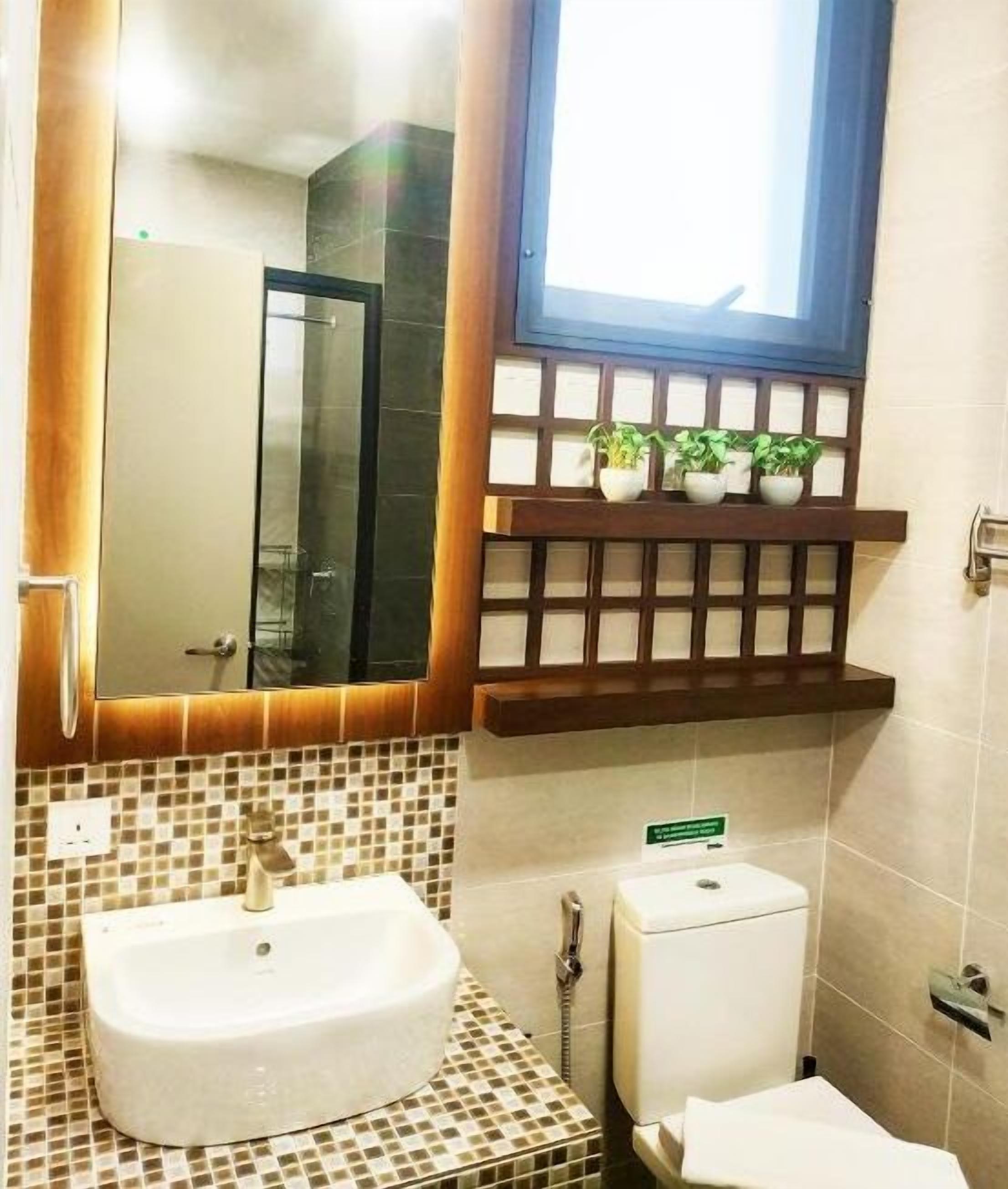 Un baño con un lavamanos blanco, un inodoro y un espejo. Hay una ventana sobre el espejo y una repisa con plantas en macetas.